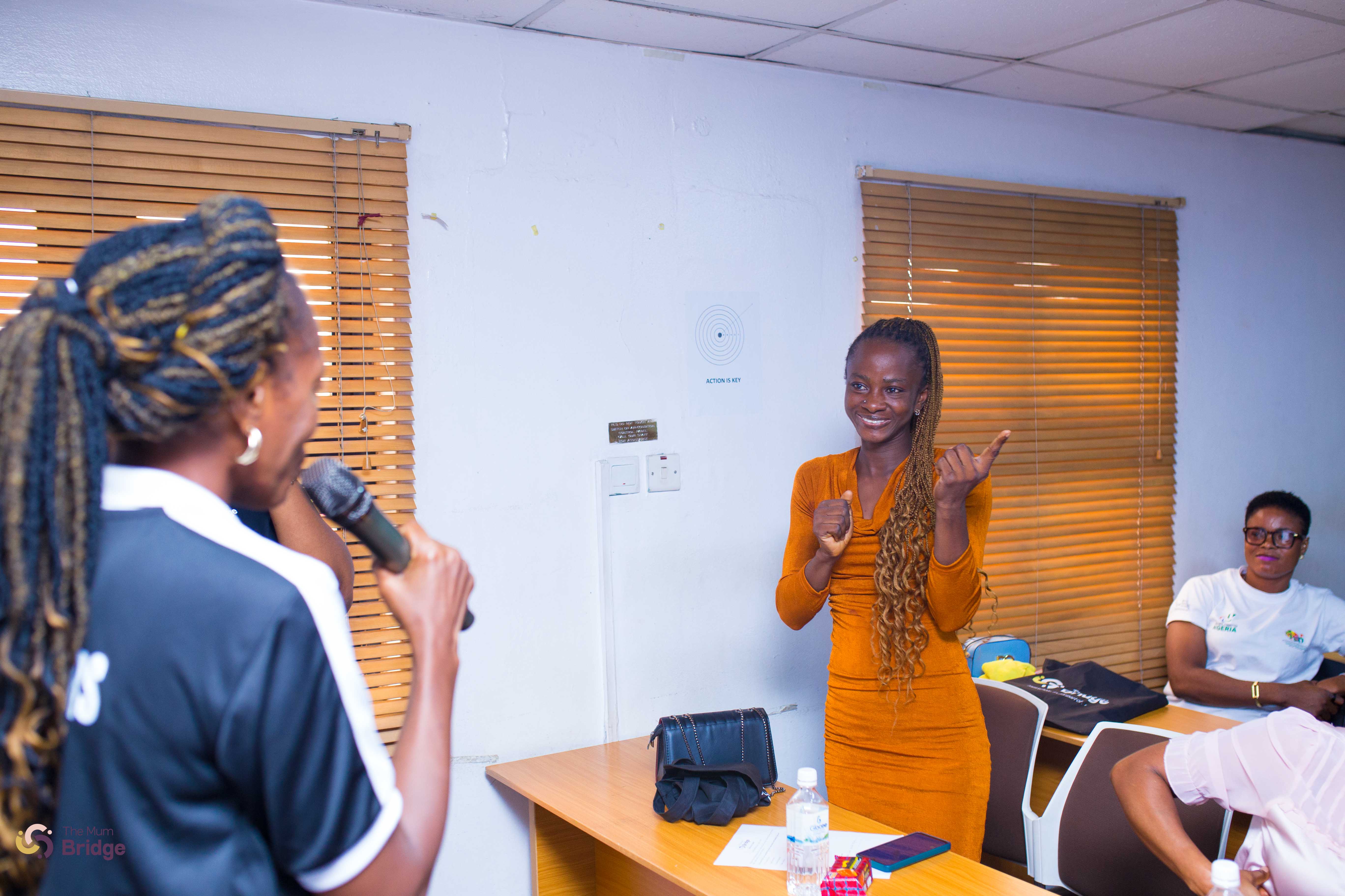 A participant in an orange dress shares her story as a Mum Bridge facilitator holds a microphone toward her