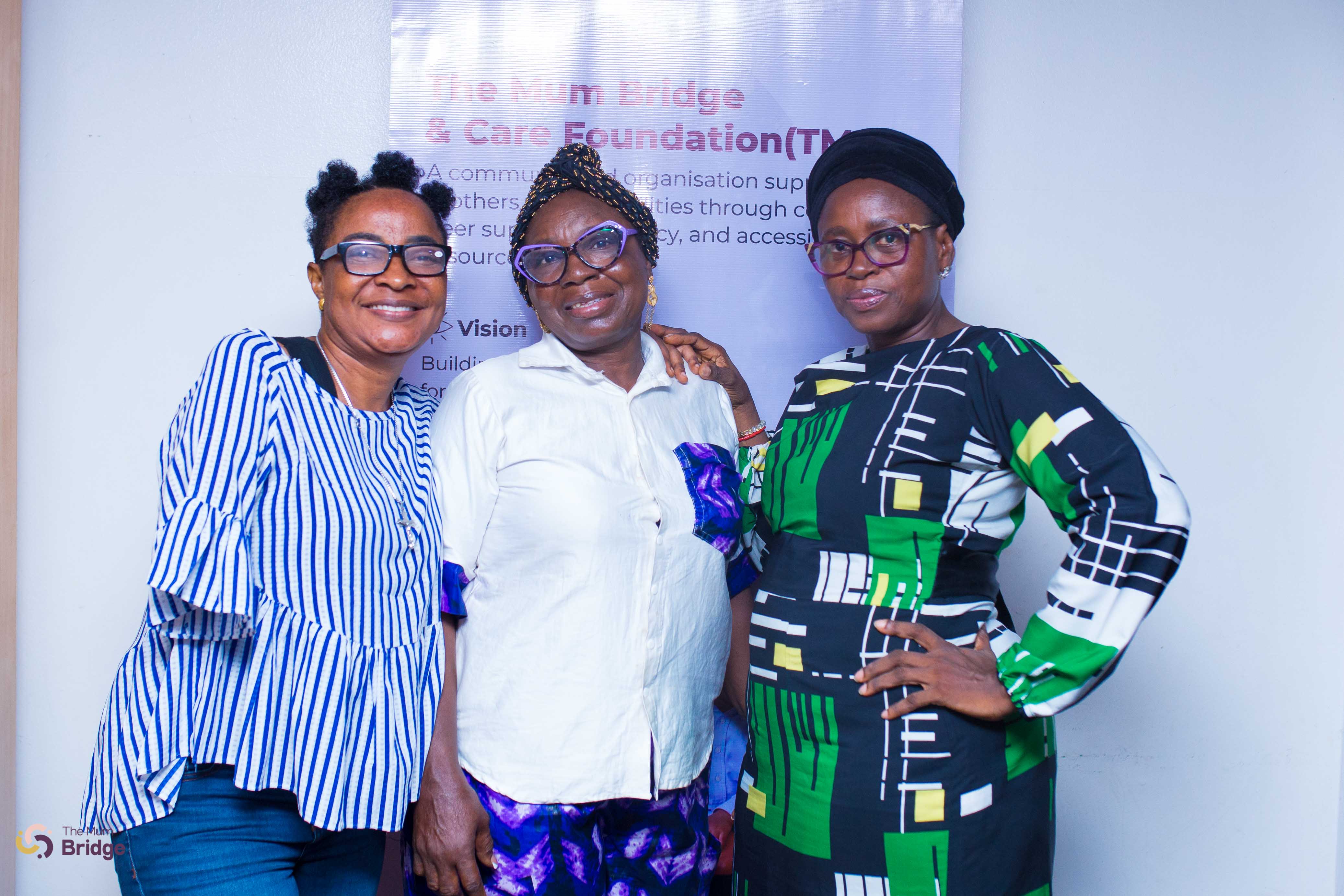 Three women smile together at a Mum Bridge community event in front of the foundation banner