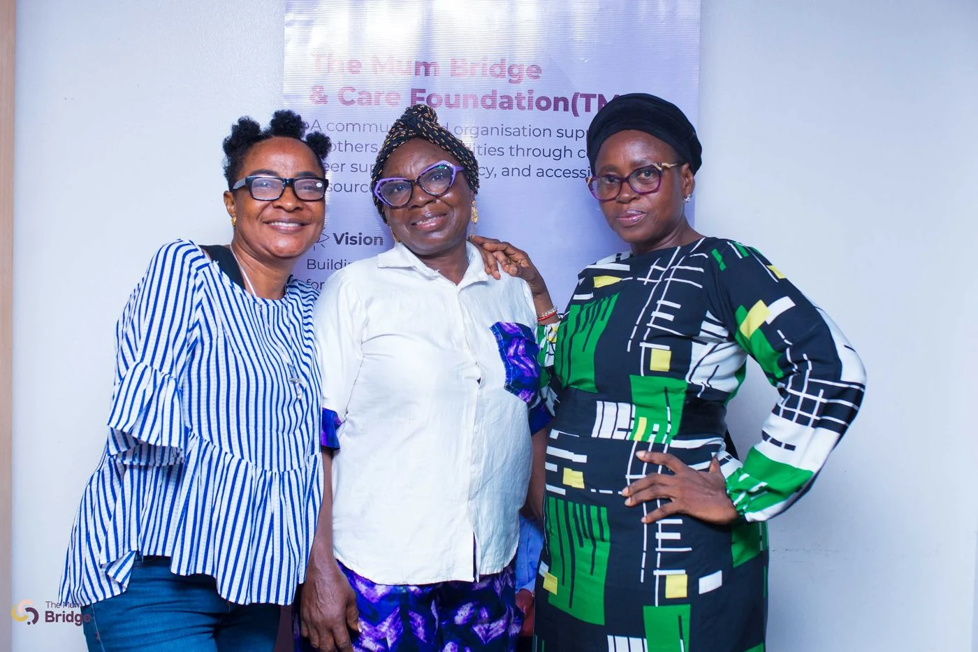 Three women smile together at a Mum Bridge community event in front of the foundation banner