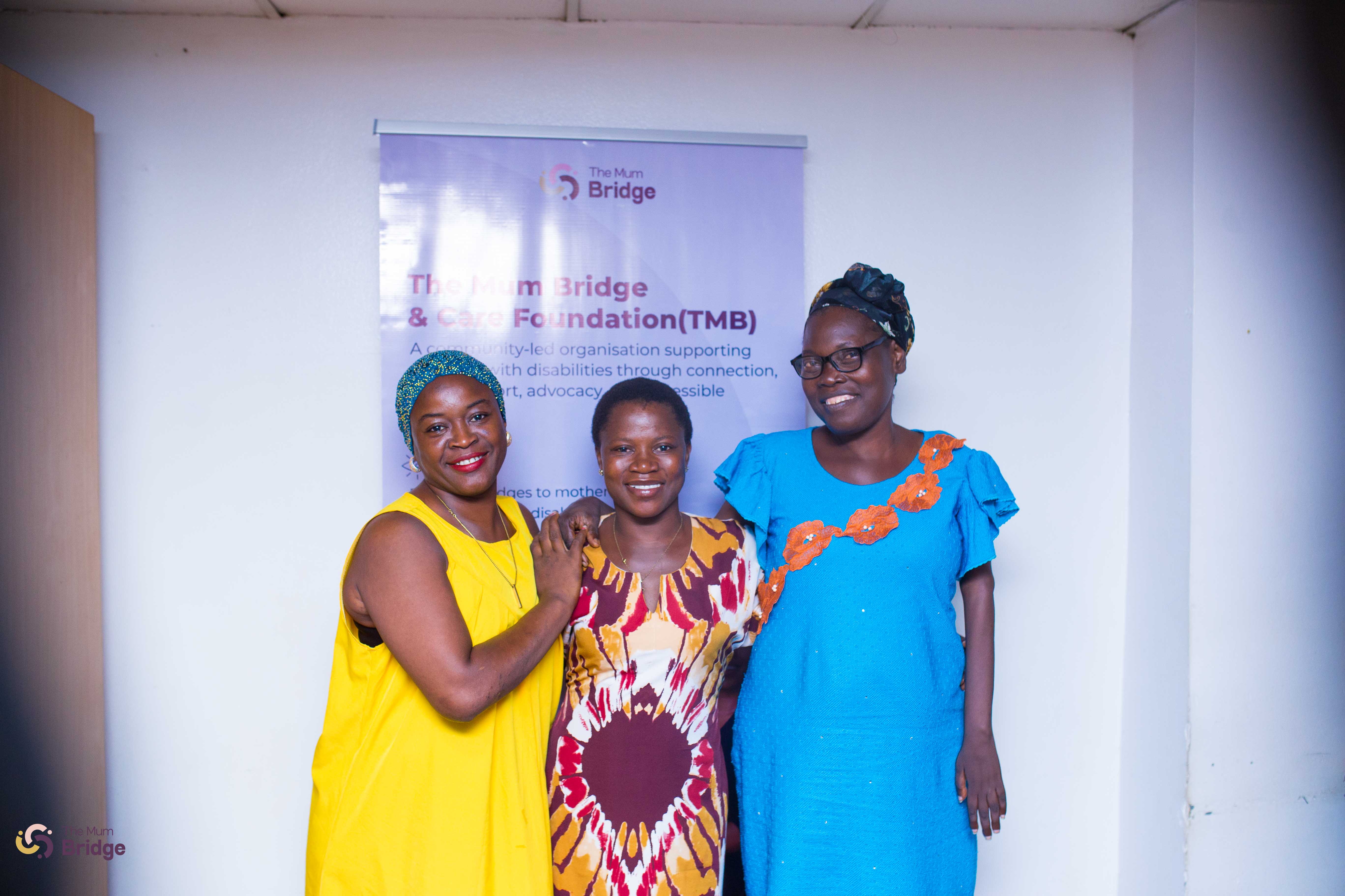 Three community members smile warmly together in front of The Mum Bridge & Care Foundation banner