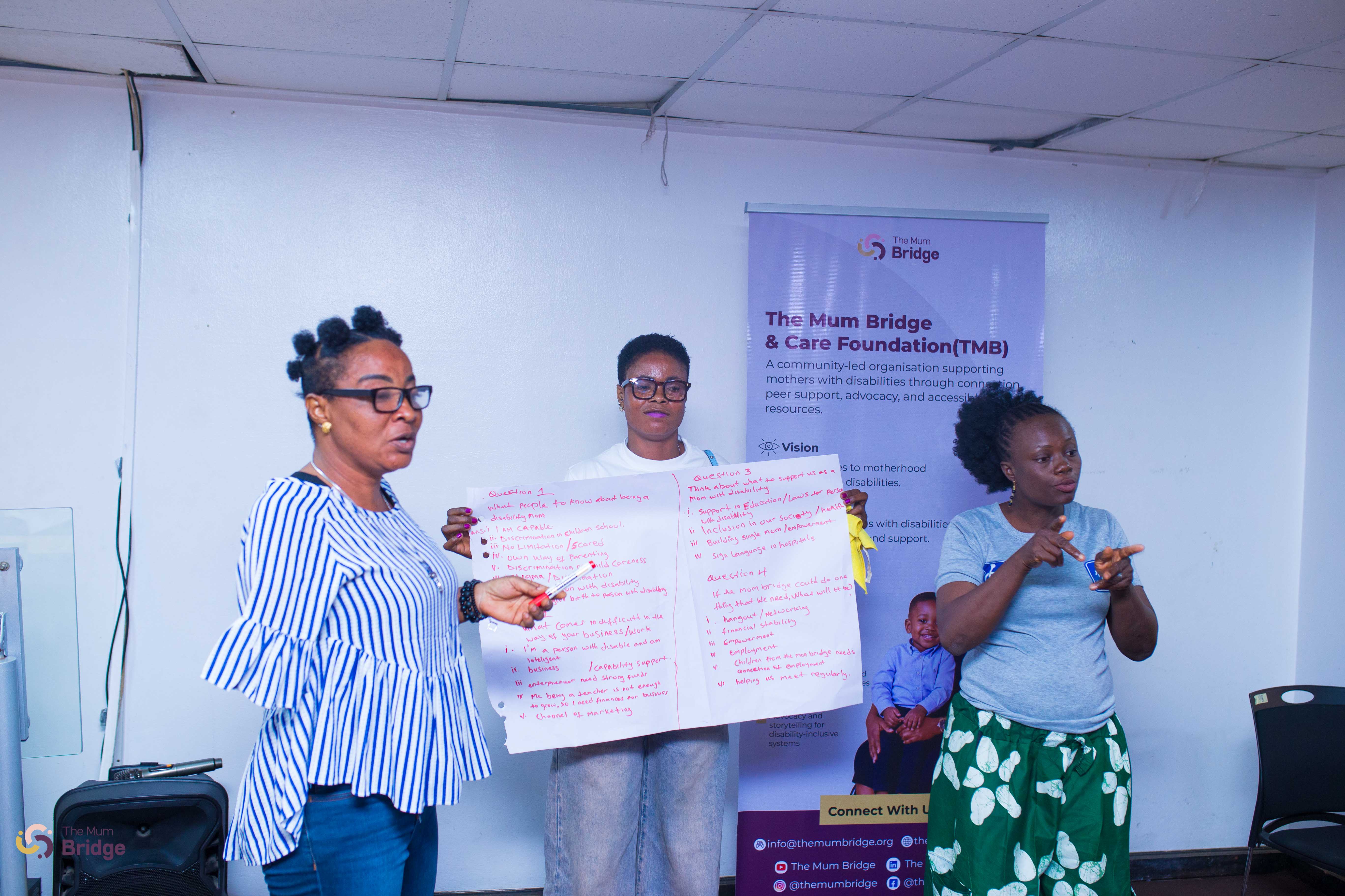 Three women present workshop findings on a flip chart in front of the Mum Bridge banner, with a sign language interpreter alongside