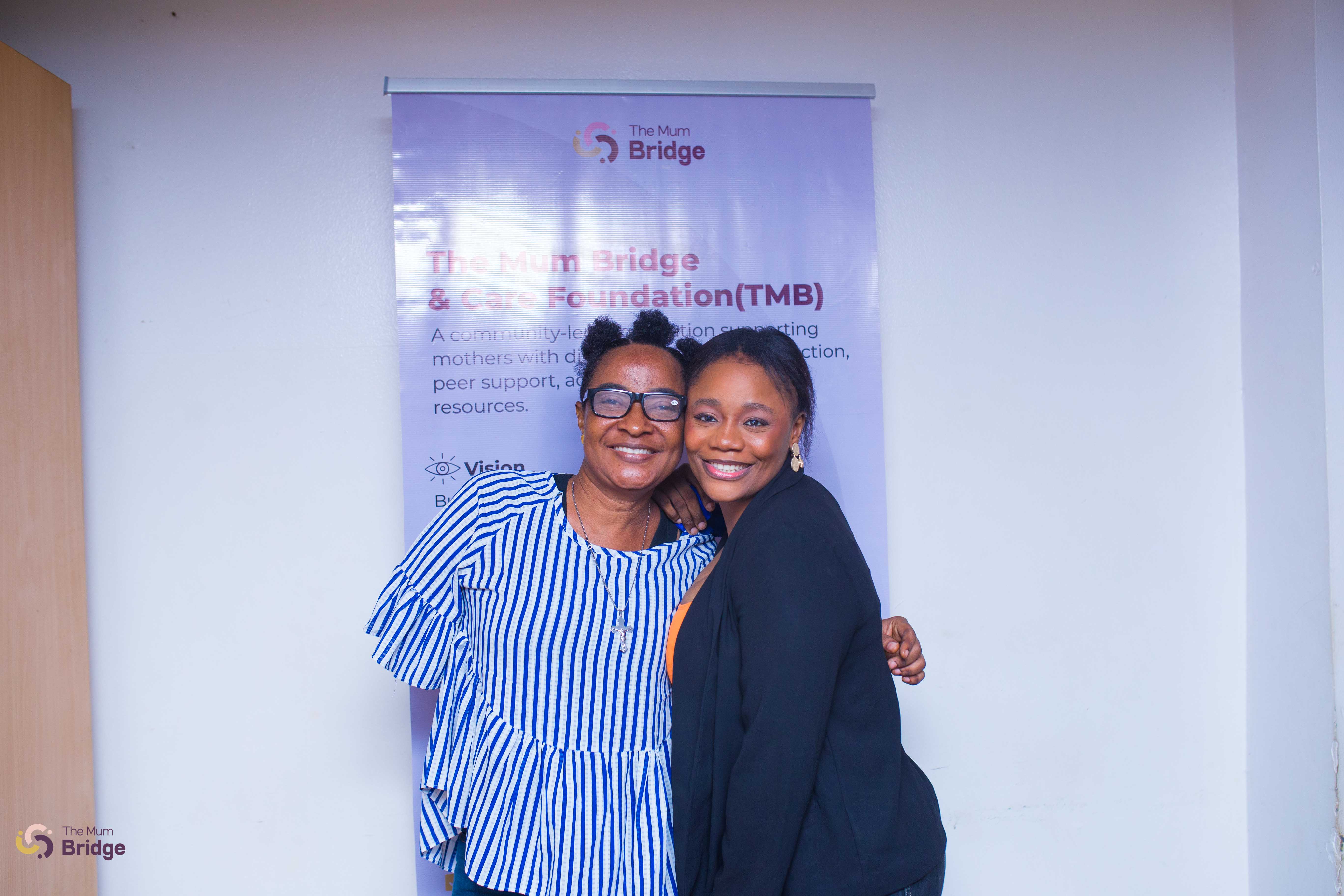Two attendees share a warm embrace in front of The Mum Bridge & Care Foundation banner