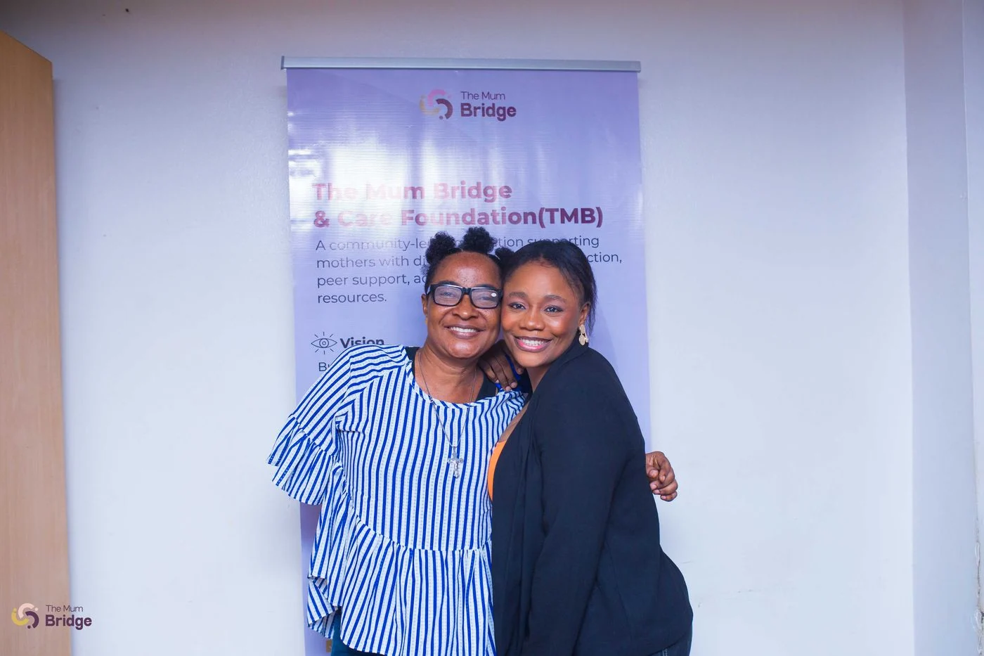 Two attendees share a warm embrace in front of The Mum Bridge & Care Foundation banner