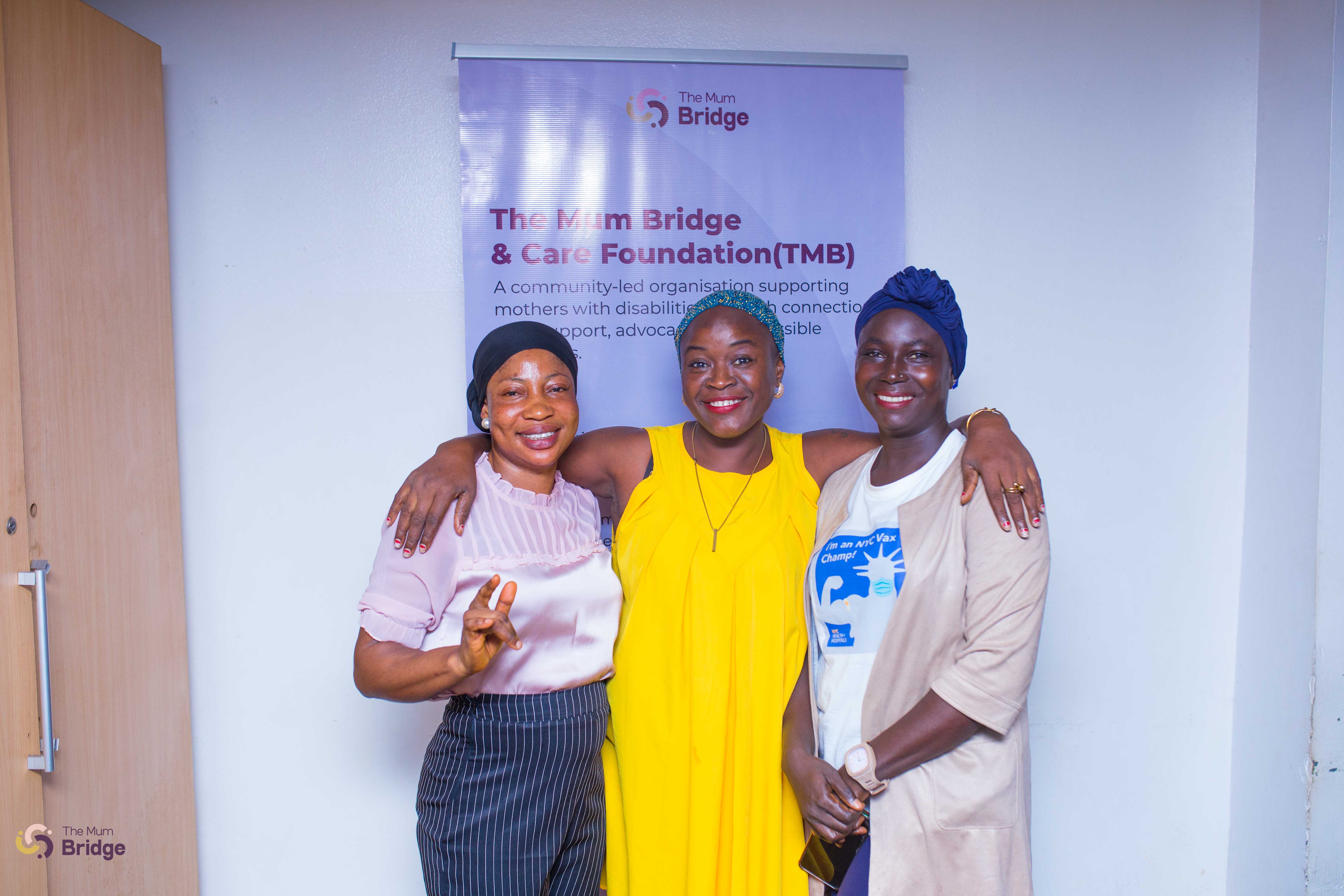 Three women pose together at a Mum Bridge event, with the foundation banner visible behind them
