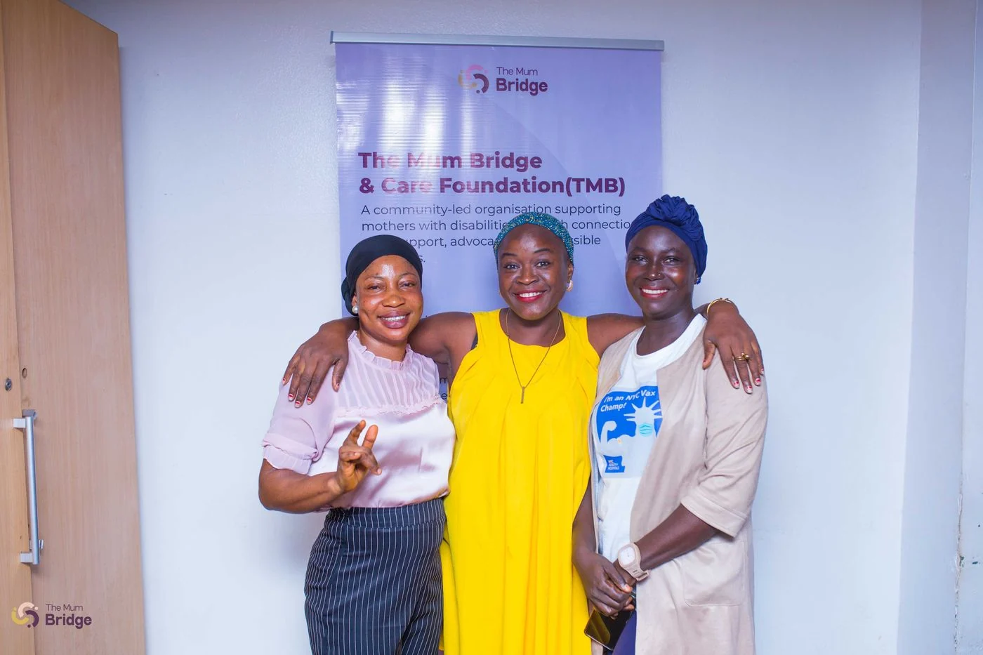 Three women pose together at a Mum Bridge event, with the foundation banner visible behind them