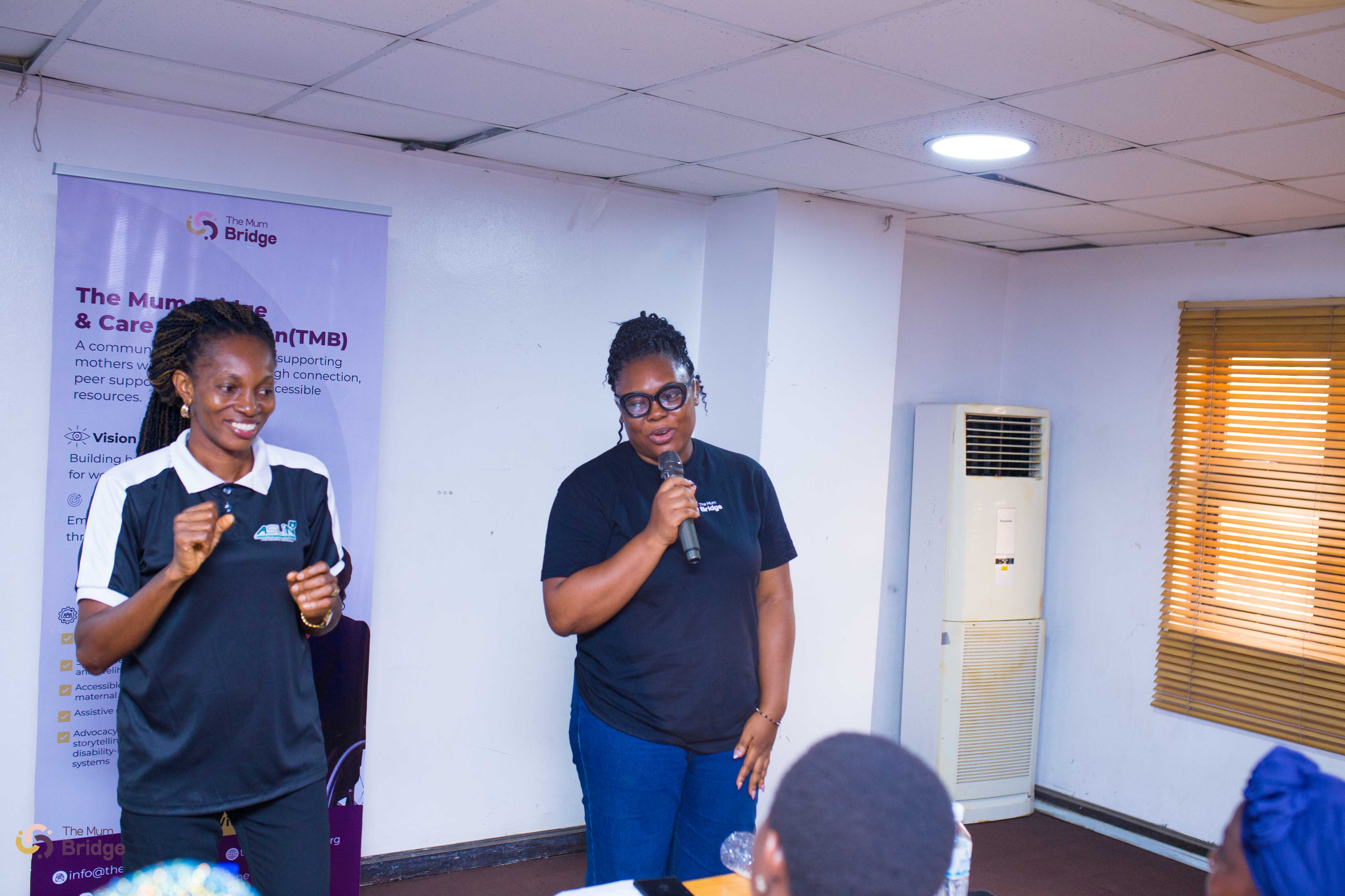 Two Mum Bridge facilitators at the front of the room, one speaking into a microphone with the foundation banner visible behind them